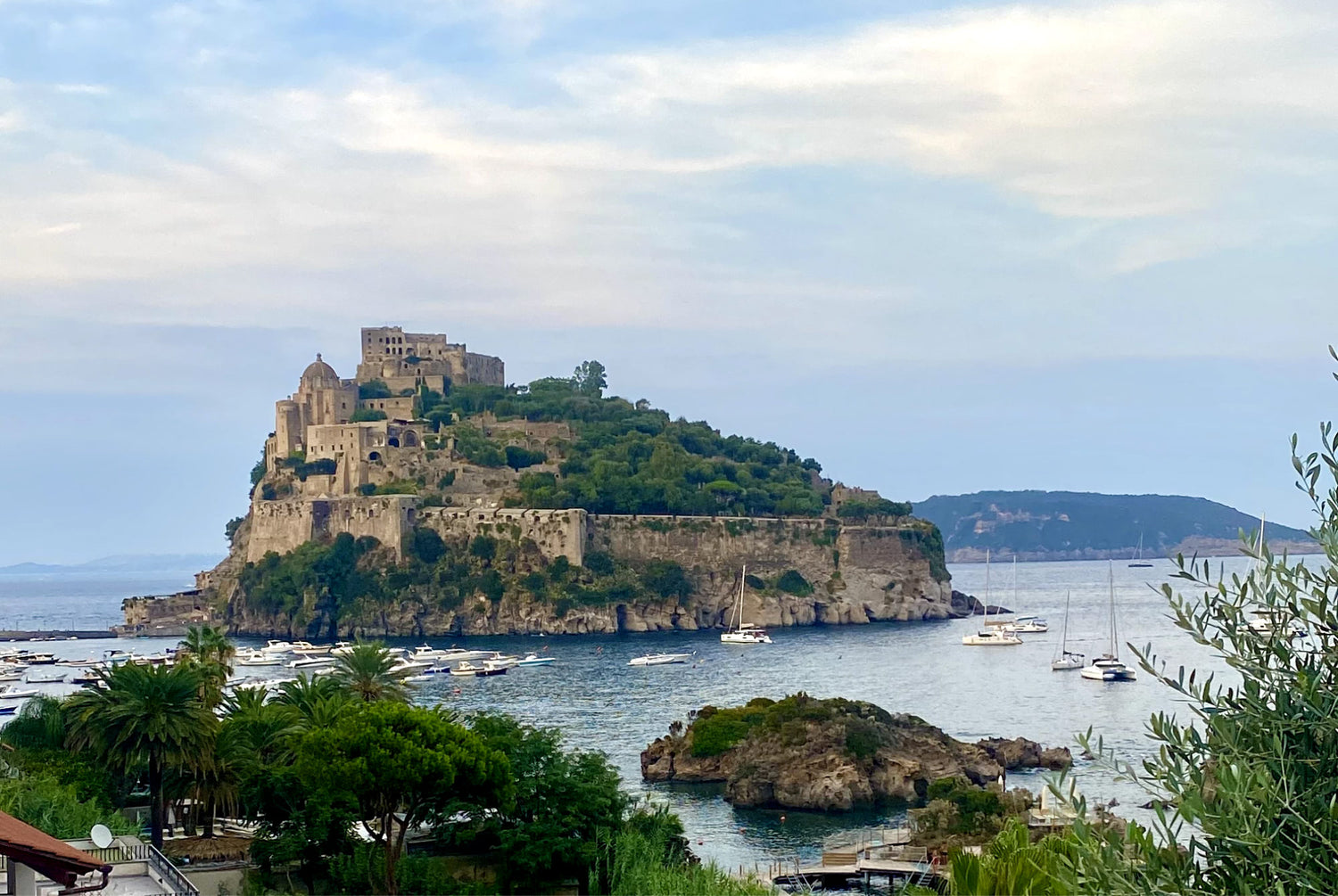 Island is Ischia, Italy with a castle and boats surrounding it, as well as blue sky and lush green plants.